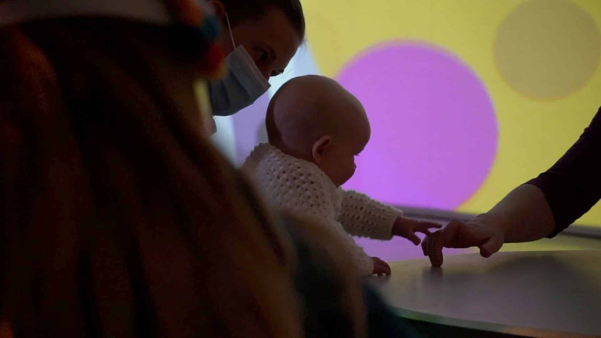 baby holding out their hand to an adult with brightly coloured shapes in the background on the wall