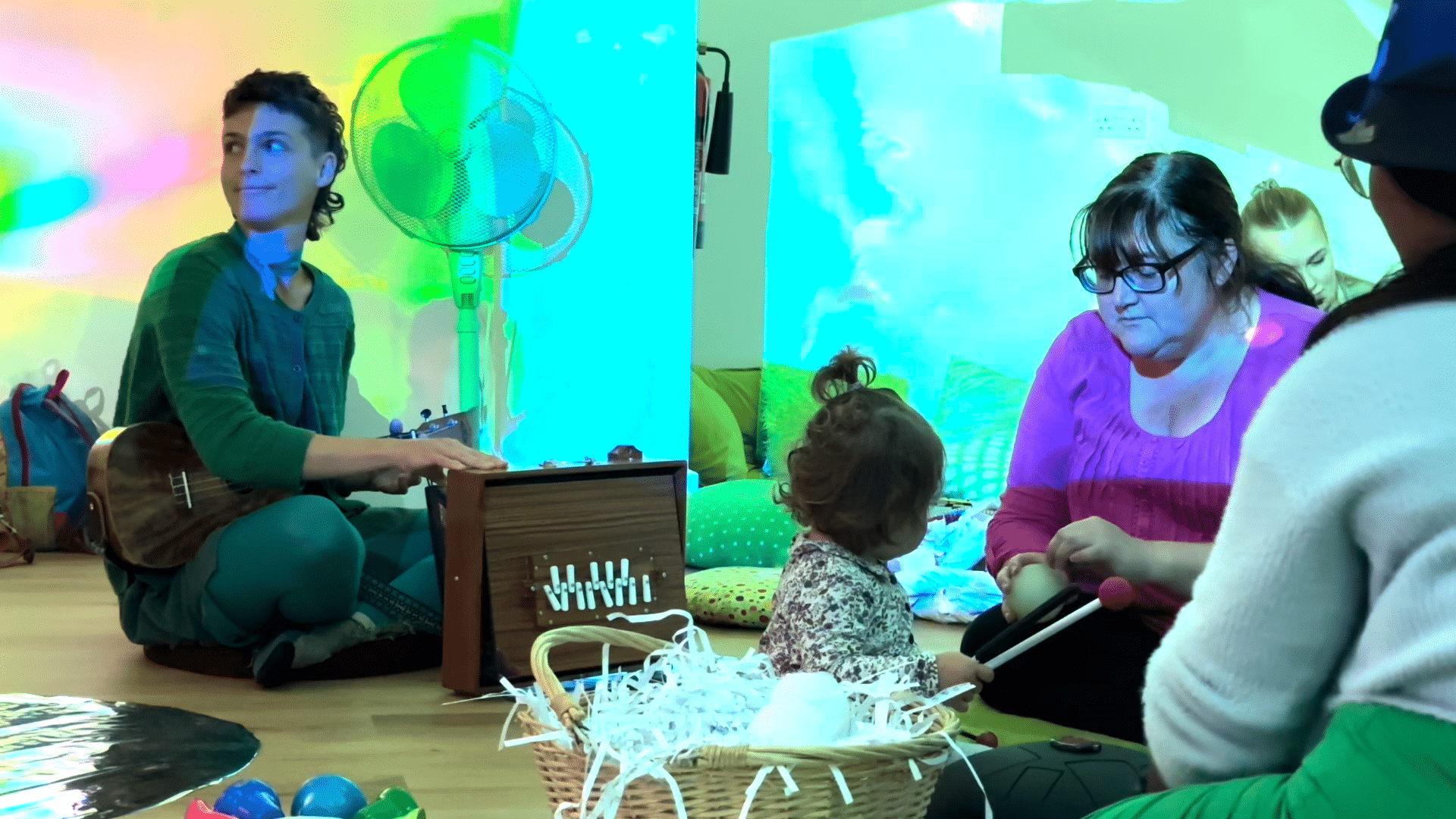 group of people sat with a child during a creative workshop, there are lots of bright coloured lights and textures on the walls
