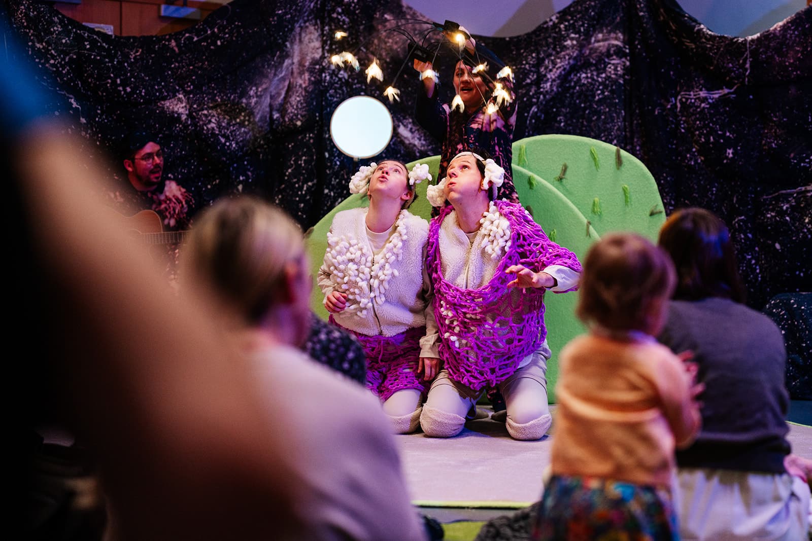 Two people on stage wearing brightly coloured costumes looking up at the ceiling